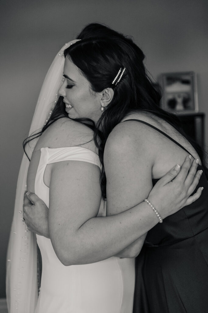 Bride in a white wedding dress embracing a bridesmaid with joy, captured in a candid black and white photo.