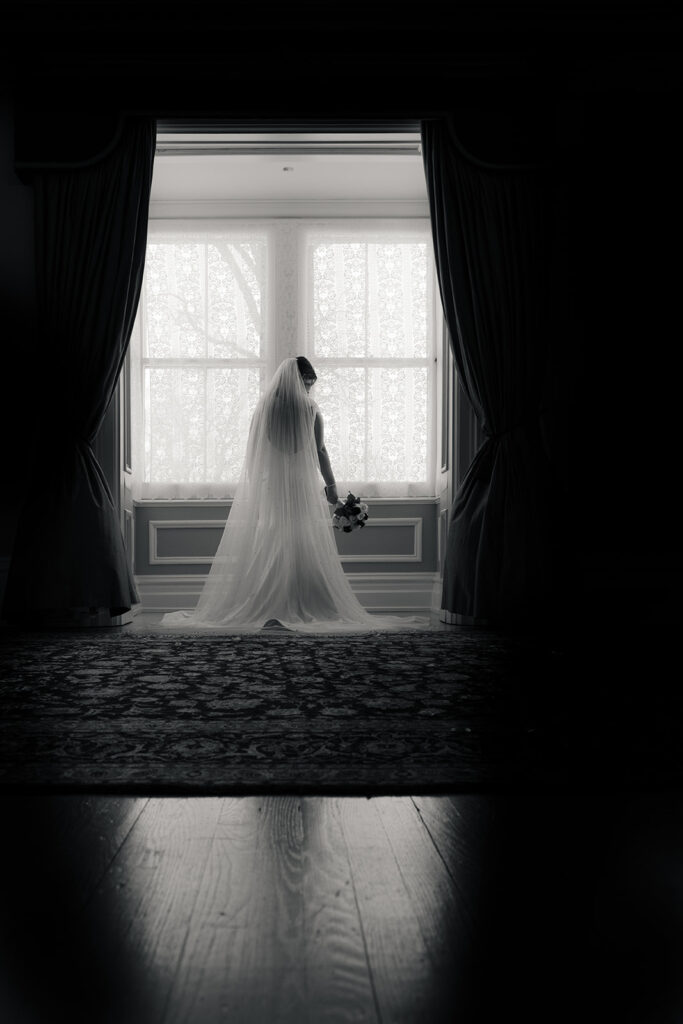 Bride in silhouette against a bright window, her veil trailing gracefully, holding a bouquet.