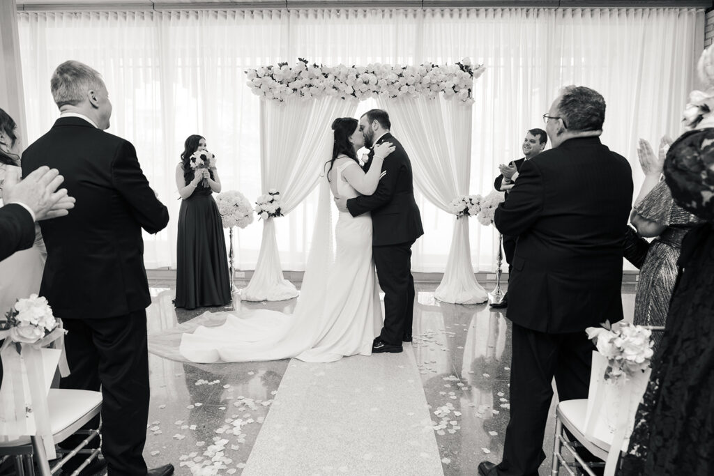 Newlyweds first kiss passionately, surrounded by applauding guests and elegant floral decorations."