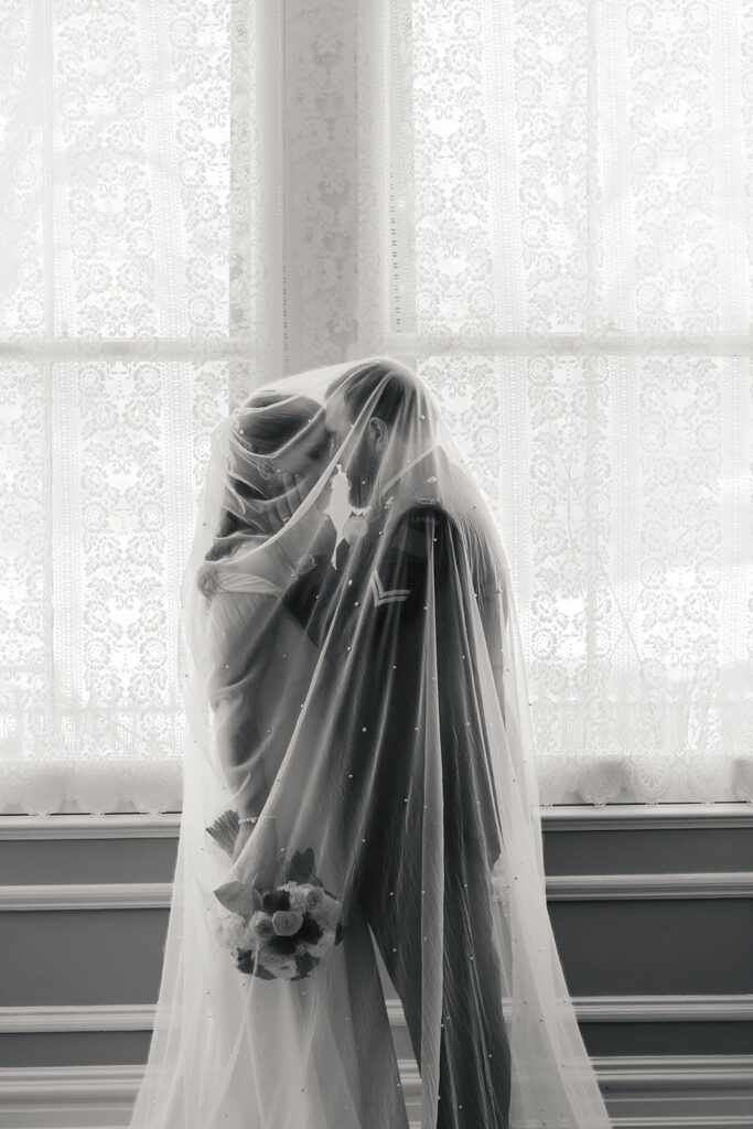Bride and groom enveloped in the bridal veil, sharing a tender kiss by the window light.