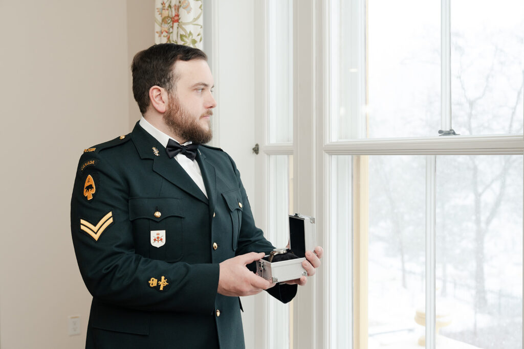 Groom in military uniform holding a box with wedding rings by a window with a snowy view