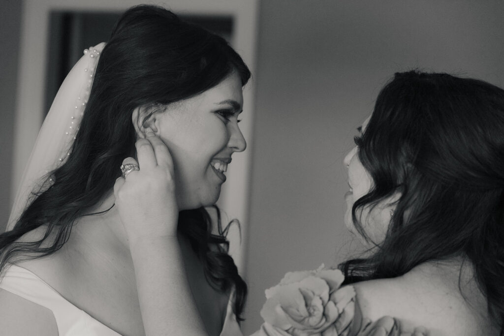 Joyful bride being assisted with her earring by a bridesmaid, sharing a smile, in a candid black and white photo.