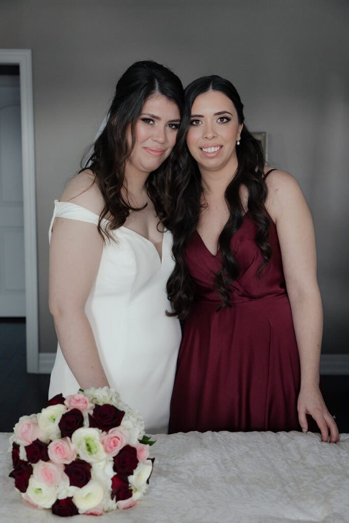 Bride in a white dress smiling with her bridesmaid in a maroon dress, with a bouquet of white and dark red roses on the table in the foreground.