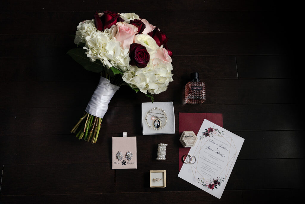 Wedding day essentials flat lay with white and dark red floral bouquet, Valentino perfume, jewelry, and invitation suite on a dark wooden background