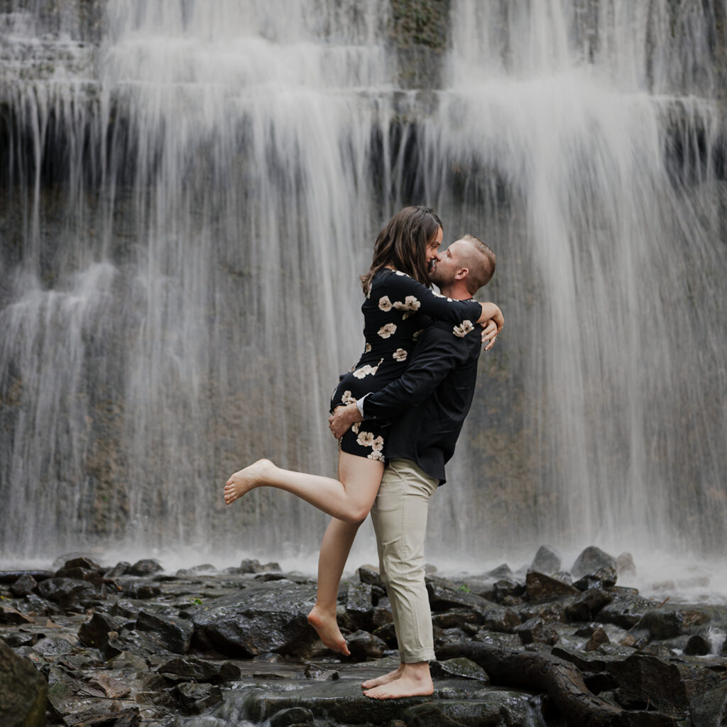 engaged couple barefoot on the wet rocks and behind them is a waterfall with rushing water