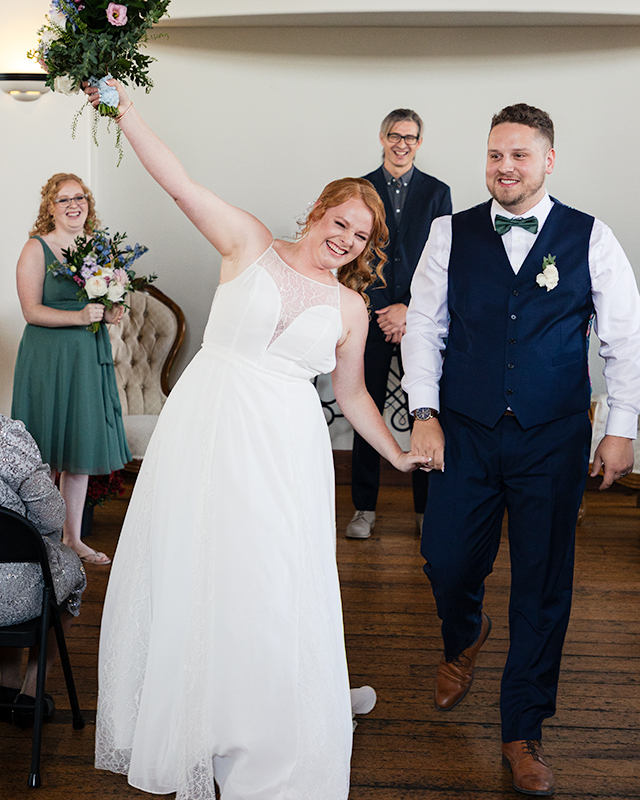 couple walking down the aisle just married—bride lifts bouquet while guests cheer, candid Toronto/KW wedding photo