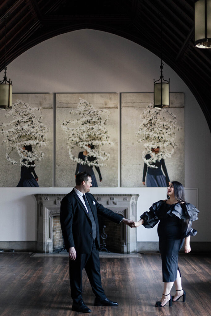 Couple cuddling by gothic windows at U of T, soft side-light—architectural engagement portrait.