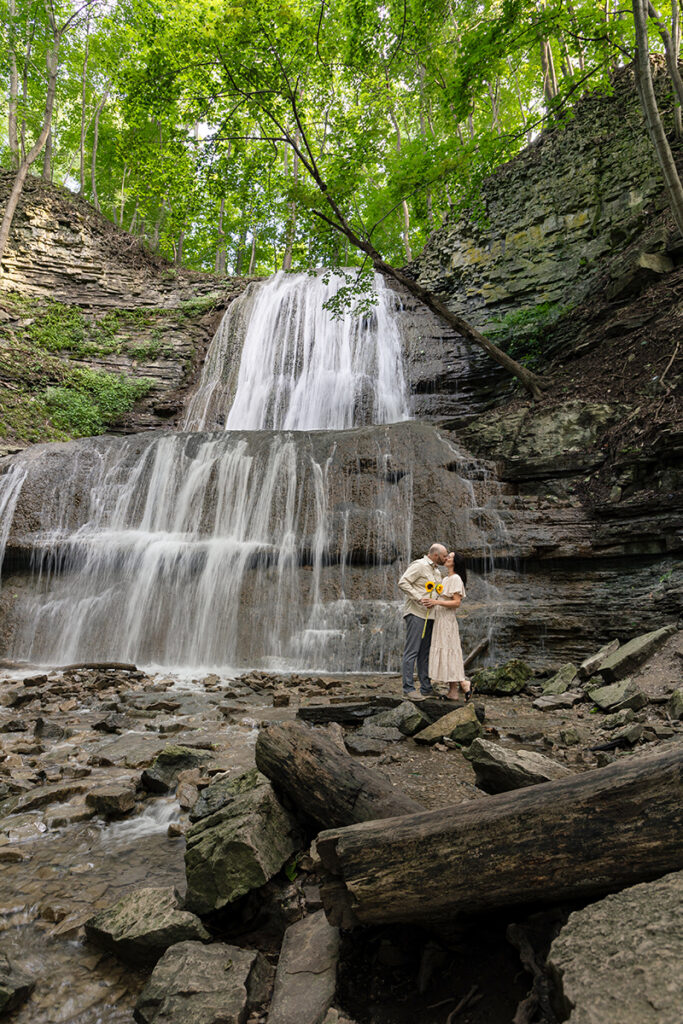 Couple kissing at the base of a forest waterfall, mist in the air—wide, cinematic frame.