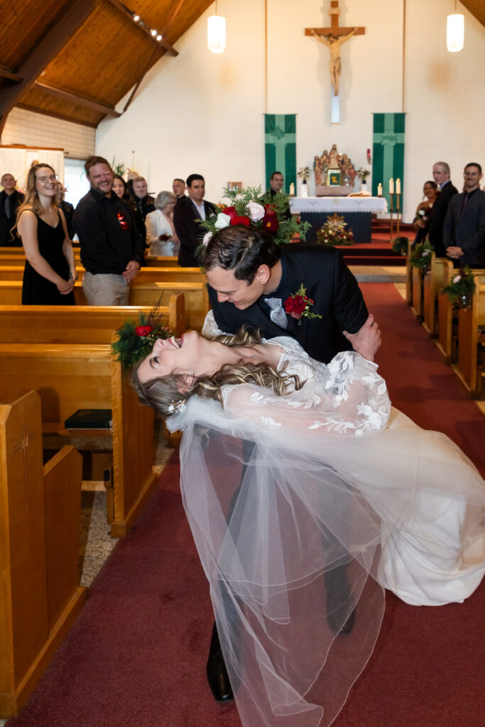 Candid wedding photo of a groom dipping his laughing bride in a church aisle, guests cheering in the background, veil sweeping.