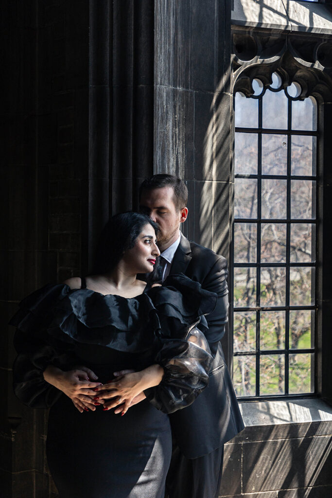 Couple cuddling by gothic windows at U of T, soft side-light—architectural engagement portrait.