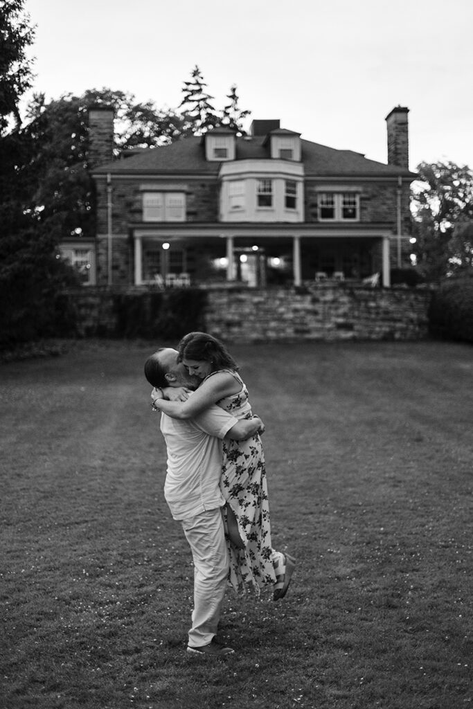 Couple hugging on Paletta Mansion lawn at dusk, soft house lights behind—elegant garden portrait.